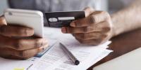 Online payment and shopping concept. Cropped shot of African-American male holding cell phone in one hand and credit card in other, making transaction, suing mobile banking app during lunch at cafe